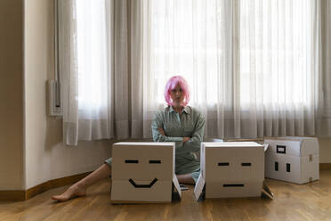 Serious young woman wearing pink wig sitting on the floor with smiley and frowning face cardboard box - AFVF06098