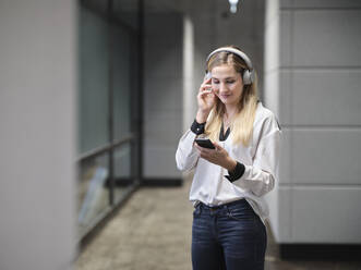 Smiling businesswoman with headphones and cell phone in modern office - CVF01579