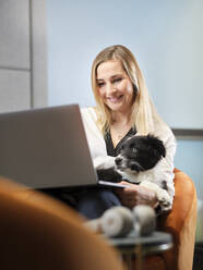 Geschäftsfrau mit Hund im Sessel sitzend mit Laptop - CVF01583