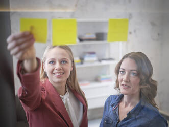 Two businesswomen working on sticky notes at glass pane in office - CVF01597