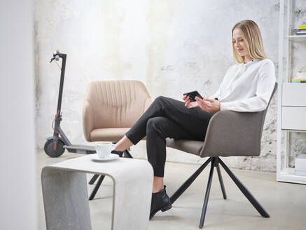 Relaxed businesswoman using cell phone in modern office with electric scooter in background - CVF01604