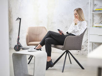 Entspannte Geschäftsfrau, die in einem modernen Büro mit einem Elektroroller im Hintergrund ein Mobiltelefon benutzt - CVF01611