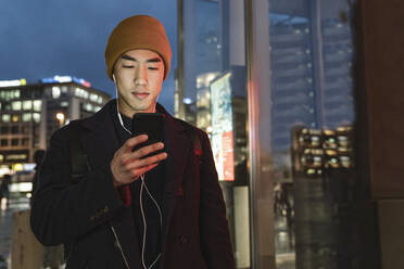 Stylish man with yellow hat and earphones using smartphone at evening on the street at night - AHSF02259