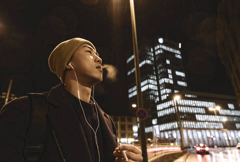 Stylish man with yellow hat and earphones smoking a cigarette in the city at night stock photo