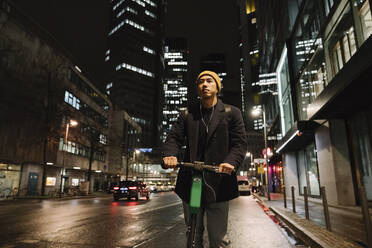 Man with yellow hat and earphones using scooter in the city at night - AHSF02276