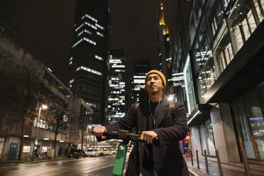 Mann mit gelbem Hut und Kopfhörern auf einem Motorroller in der Stadt bei Nacht - AHSF02277