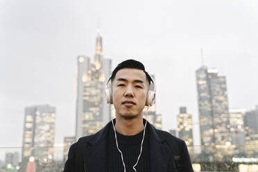 Porträt eines Mannes mit Kopfhörern vor einer städtischen Skyline, Frankfurt, Deutschland - AHSF02286