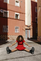 Smiling young woman wearing red dress and sitting on ground, stretching her legs - TCEF00506