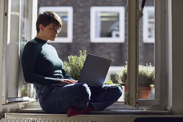 Woman sitting at the window at home using laptop - MCF00668