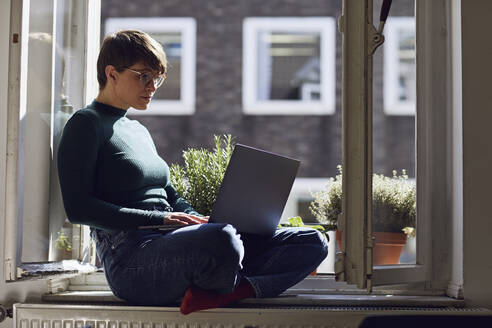 Frau sitzt zu Hause am Fenster und benutzt einen Laptop - MCF00668