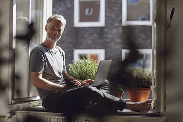 Mature man sitting at the window at home using laptop - MCF00669