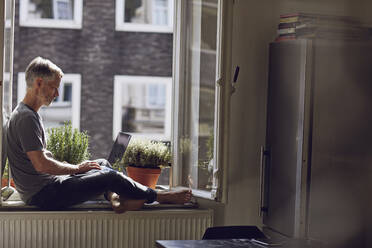 Mature man sitting at the window at home using laptop - MCF00670