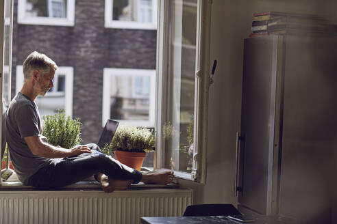 Mature man sitting at the window at home using laptop - MCF00670