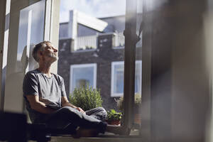 Älterer Mann sitzt mit geschlossenen Augen am Fenster zu Hause - MCF00671