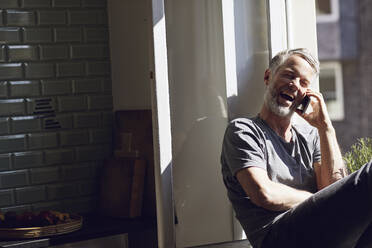 Laughing mature man sitting at the window at home talking on the phone - MCF00676