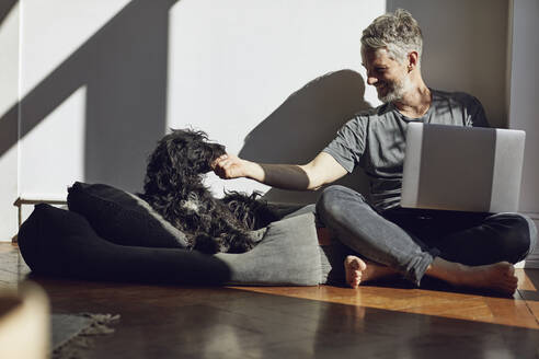 Älterer Mann sitzt zu Hause mit Laptop und Hund auf dem Boden - MCF00677