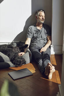 Mature man sitting on the floor at home with dog - MCF00679