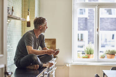 Mature man with cell phone sitting on kitchen counter at home - MCF00682