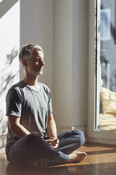 Mature man sitting on the floor at home meditating - MCF00690