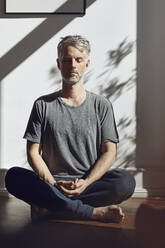 Mature man sitting on the floor at home meditating - MCF00691