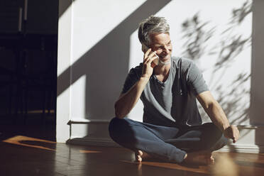 Smiling mature man sitting on the floor at home talking on the phone - MCF00693