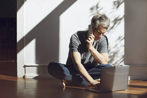Älterer Mann sitzt zu Hause auf dem Boden und benutzt Laptop und Handy - MCF00694