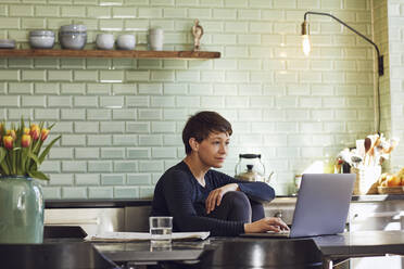 Woman sitting at tkitchen table at home using laptop - MCF00704