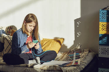 Mädchen sitzt zu Hause auf dem Bett, macht Hausaufgaben und benutzt ein Smartphone - MCF00705