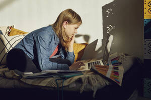 Mädchen sitzt zu Hause auf dem Bett, macht Hausaufgaben und benutzt einen Laptop - MCF00706