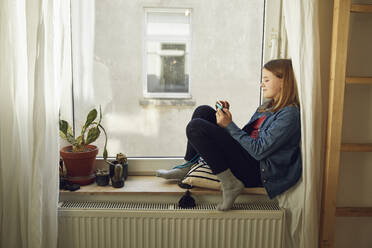 Mädchen sitzt zu Hause am Fenster und benutzt ein Smartphone - MCF00711