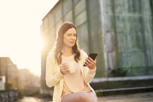 Businesswoman looking at mobile phone while having coffee on terrace - MMIF00199