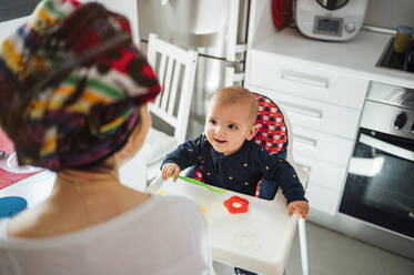 Porträt eines kleinen Jungen auf einem Hochstuhl in der Küche mit seiner Mutter - JCMF00590