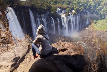 Back view of woman sitting on a rock looking at Kalandula Falls, Angola - VEGF01940