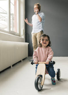 Crying girl on balance bicycle with father on the phone in background - JOSEF00283