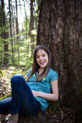 Portrait of laughing girl leaning against tree trunk in forest - LVF08840