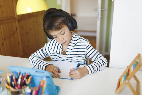 Boy doing homeschooling and writing on notebook, using tablet and headphones at home - HMEF00898
