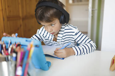 Boy doing homeschooling and writing on notebook, using tablet and headphones at home - HMEF00899