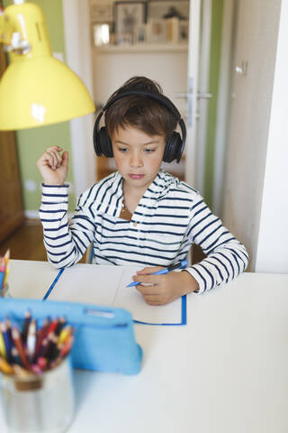 Junge, der zu Hause Hausaufgaben macht und auf einem Notebook schreibt, Tablet und Kopfhörer benutzt, lizenzfreies Stockfoto