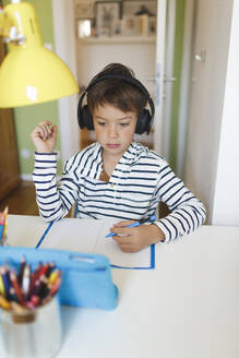 Boy doing homeschooling and writing on notebook, using tablet and headphones at home - HMEF00900