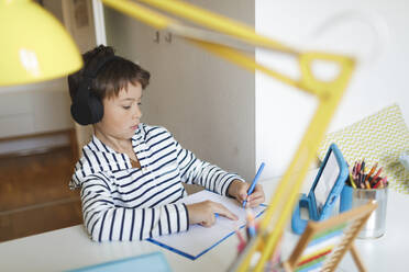 Boy doing homeschooling and writing on notebook, using tablet and headphones at home - HMEF00901