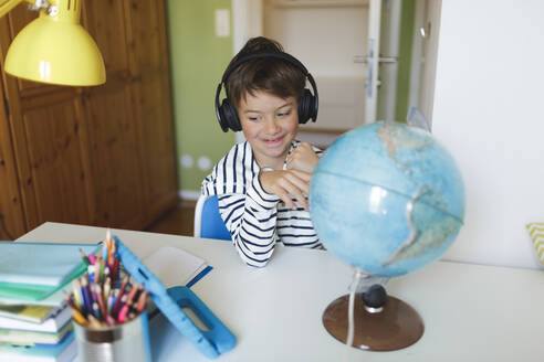 Boy doing homeschooling with a globe and using tablet and headphones at home - HMEF00903