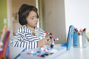 Boy doing homeschooling and holding molecule model, using tablet and headphones at home - HMEF00904