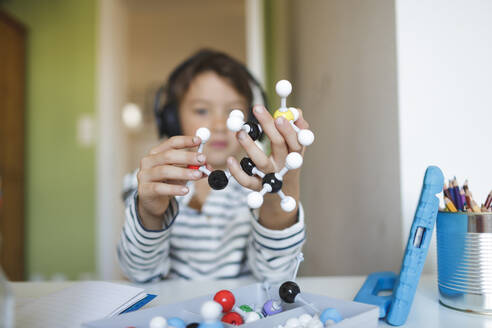 Boy doing homeschooling and holding molecule model, using tablet and headphones at home - HMEF00905