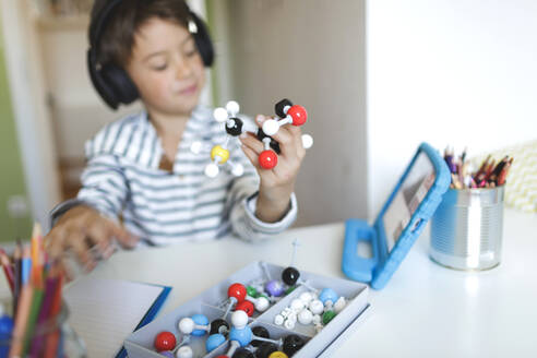 Boy doing homeschooling and holding molecule model, using tablet and headphones at home - HMEF00906