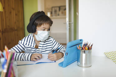 Junge beim Hausunterricht und beim Schreiben auf einem Notebook, mit Tablet und Kopfhörern, mit Maske zu Hause während der Corona-Krise - HMEF00907