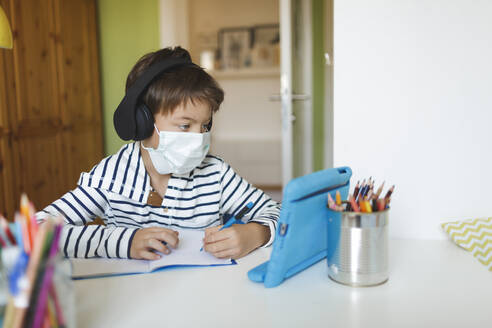 Boy doing homeschooling and writing on notebook, using tablet and headphones, wearing mask at home during corona crisis - HMEF00907