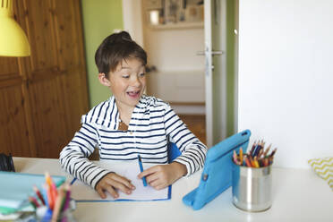 Boy doing homeschooling and writing on notebook, using tablet and headphones at home - HMEF00908