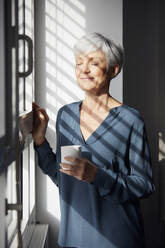 Porträt einer älteren Geschäftsfrau mit Kaffeetasse, die am Fenster steht und das Sonnenlicht genießt - RBF07568