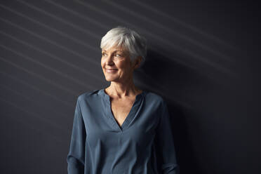 Portrait of smiling senior woman against grey wall looking at distance - RBF07569