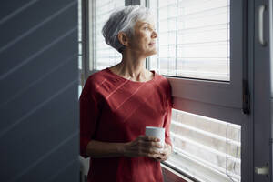 Portrait of senior woman with coffee mug looking out of window - RBF07607